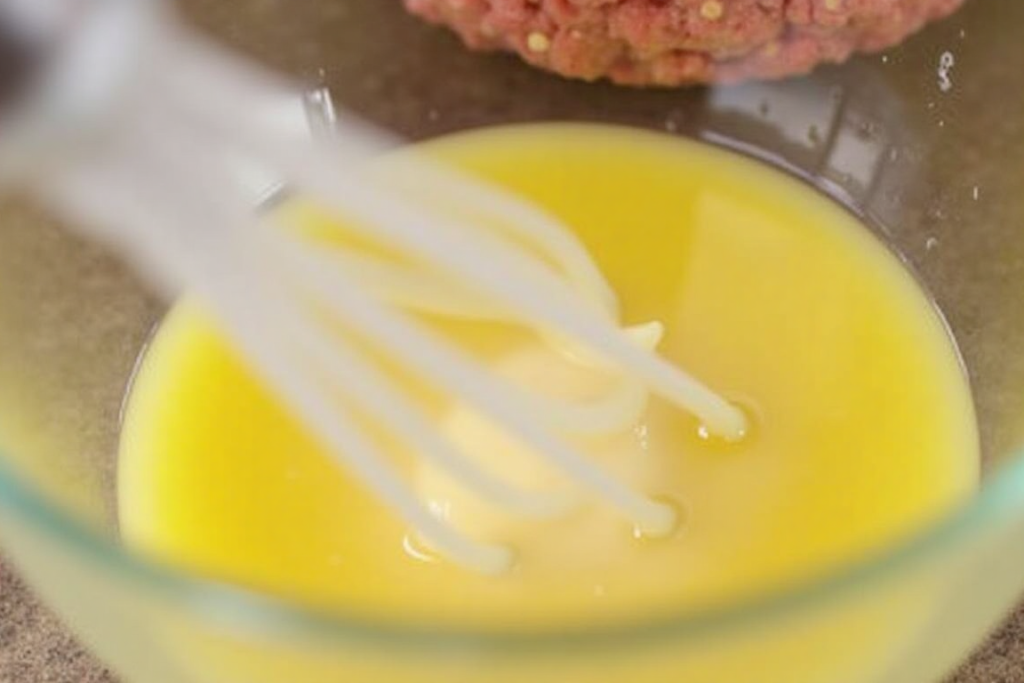 Whisked egg whites in a bowl with burger patty mixture nearby.