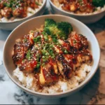 Close-up of a sticky chicken rice bowl topped with spicy mayo, sesame seeds, green onions, and served with broccoli